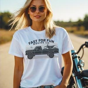 以下が含まれることがあります： 黒と白のピックアップトラックにモトクロスバイクを積んだグラフィックがプリントされた白いTシャツ。Tシャツには「FAST FOR FUN MOVING CO. EST. 2018」の文字がプリントされています。