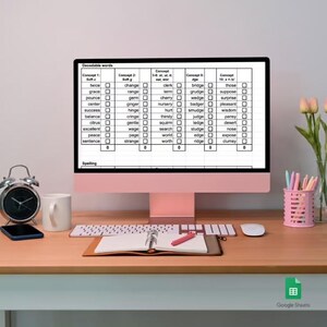 Pode incluir: Um monitor de computador exibe uma planilha intitulada Decodable words com colunas para conceitos como Soft c, Soft g e dge. A tela mostra listas de palavras com caixas de seleção. Um teclado, mouse, caderno, caneta, relógio e flores estão na mesa.