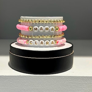 May include: A set of three bracelets with pink and white beads and gold accents. The bracelets spell out the names "MAMA" and "CLARA".
