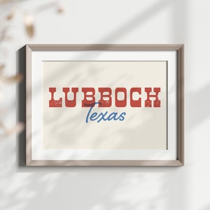 May include: A framed print displaying the word "Lubbock" in distressed red lettering above the word "Texas" in blue script. The artwork has a cream background and is set within a light brown frame.