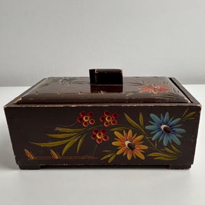 Puede incluir: Una caja rectangular de madera de color marrón oscuro con tapa y un pequeño asa. El exterior presenta motivos florales pintados a mano en rojo, naranja, amarillo y azul, acompañados de hojas y tallos verdes. Los bordes muestran signos de desgaste.