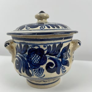 May include: A small, white ceramic pot with a lid, decorated with blue floral patterns. The pot has two handles and a knob on the lid.