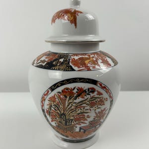 Puede incluir: Jarrón de porcelana blanca con tapa, decorado con motivos florales dorados y rojos. El jarrón tiene un diseño chino tradicional.