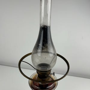 May include: An antique oil lamp with a clear glass chimney and a bulbous glass reservoir. The lamp has a metal frame and a circular handle. The base is a dark gray color. The reservoir appears to contain a reddish-brown liquid.