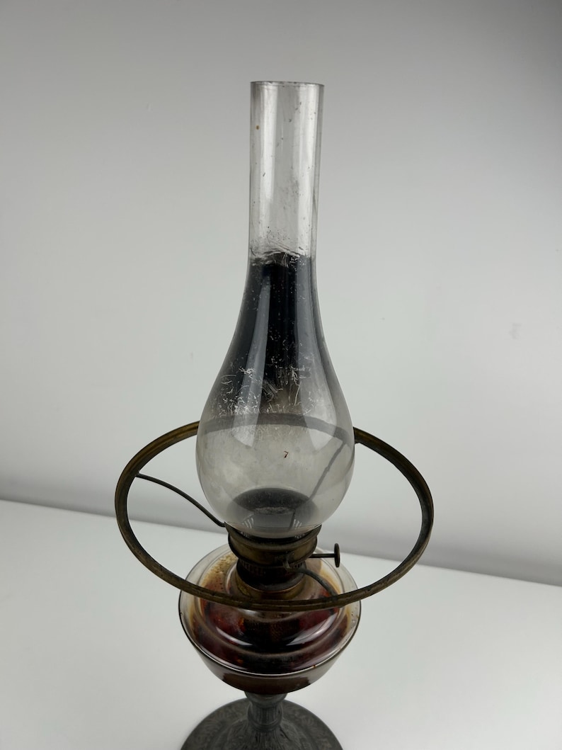 May include: An antique oil lamp with a clear glass chimney and a bulbous glass reservoir. The lamp has a metal frame and a circular handle. The base is a dark gray color. The reservoir appears to contain a reddish-brown liquid.