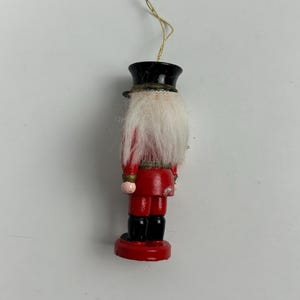 May include: A wooden Christmas ornament in the shape of a nutcracker. The ornament has a red coat, black boots, and a black hat. It features a white beard and a gold string for hanging. The ornament is approximately 4 inches tall.