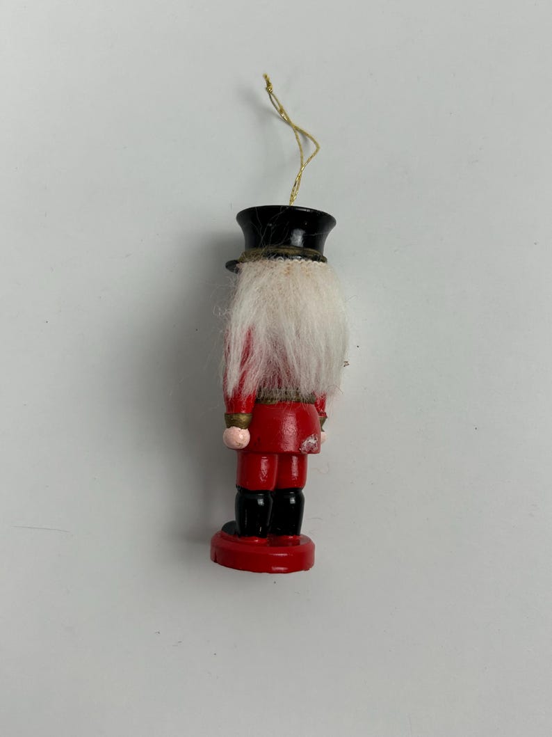May include: A wooden Christmas ornament in the shape of a nutcracker. The ornament has a red coat, black boots, and a black hat. It features a white beard and a gold string for hanging. The ornament is approximately 4 inches tall.