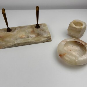 Op de afbeelding: Een set onyx bureau-accessoires, waaronder een penhouder met twee pennen, een kleine zeshoekige beker en een ovale asbak. Het onyx heeft een mix van beige, bruine en witte tinten. De pennen hebben goudkleurige accenten.