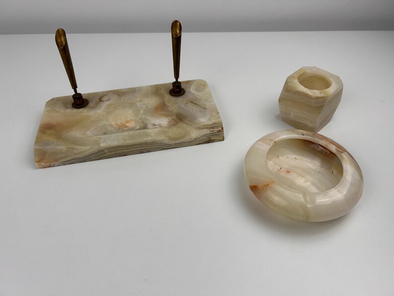 Op de afbeelding: Een set onyx bureau-accessoires, waaronder een penhouder met twee pennen, een kleine zeshoekige beker en een ovale asbak. Het onyx heeft een mix van beige, bruine en witte tinten. De pennen hebben goudkleurige accenten.