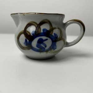 Könnte beinhalten: Eine kleine Keramik-Sahnekännchen mit rundem Körper und Henkel. Das Kännchen ist cremefarben mit braunen und blauen abstrakten floralen Designs. Der Rand und der Henkel sind braun getrimmt.
