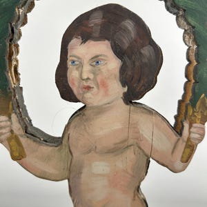 May include: A hand-painted wooden cherub figure with brown hair and blue eyes, holding golden objects. The cherub is framed by a green and gold decorative border. The figure has a distressed, antique appearance.