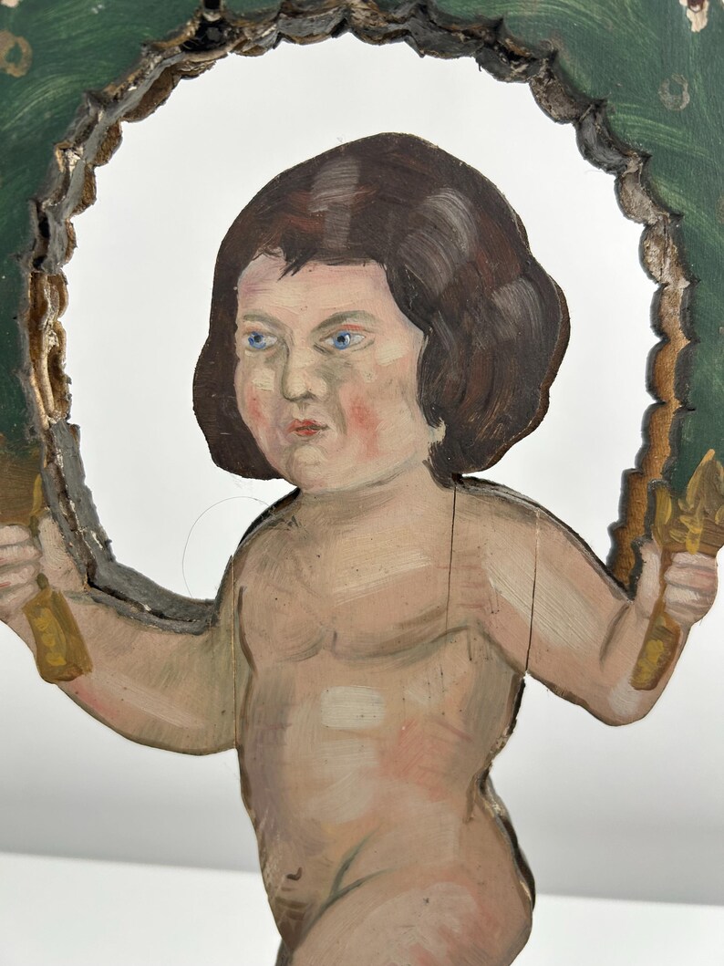 May include: A hand-painted wooden cherub figure with brown hair and blue eyes, holding golden objects. The cherub is framed by a green and gold decorative border. The figure has a distressed, antique appearance.