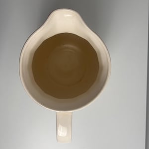 May include: A top-down view of a cream-coloured ceramic jug featuring a wide, rounded opening with a distinct pouring spout. The interior of the vessel is smooth and uniform in colour, while a single handle extends from the base of the rim.
