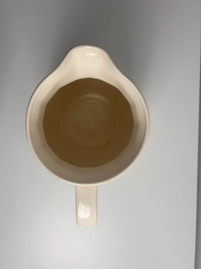 May include: A top-down view of a cream-coloured ceramic jug featuring a wide, rounded opening with a distinct pouring spout. The interior of the vessel is smooth and uniform in colour, while a single handle extends from the base of the rim.