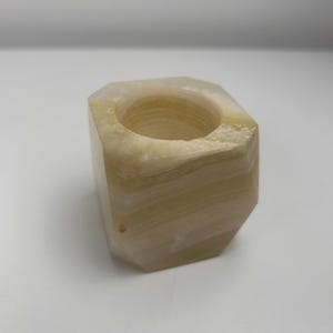 Op de afbeelding: Een natuurstenen kandelaar met een geometrisch ontwerp. De kandelaar is lichtbeige van kleur met subtiele kleurschakeringen. De binnenkant heeft een ronde opening voor een kaars. De algehele vorm is een kubus met afgeschuinde randen.