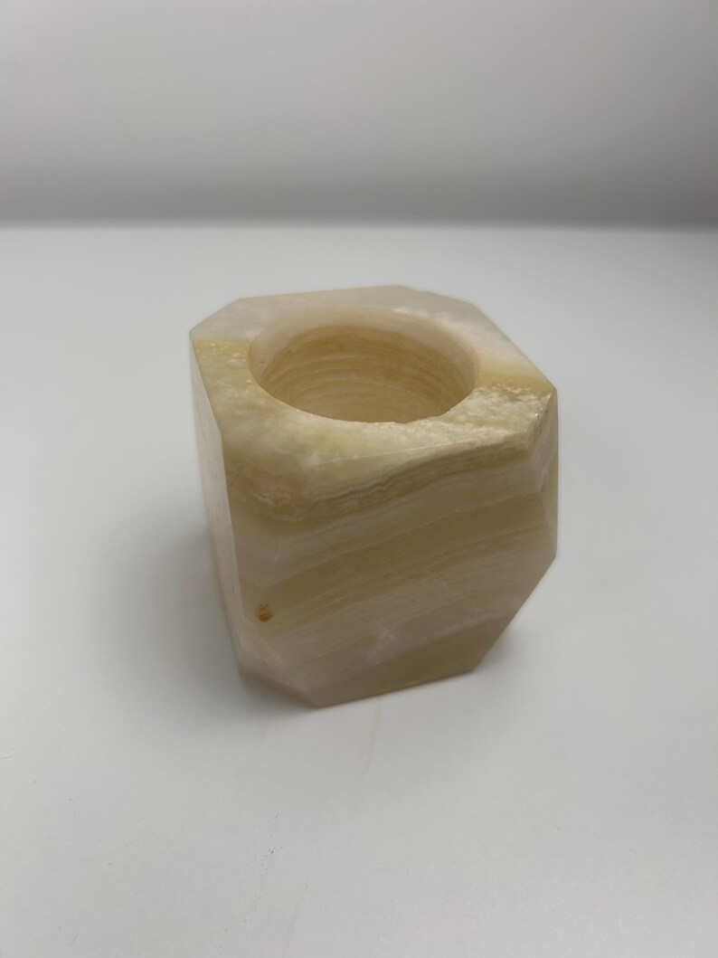 Op de afbeelding: Een natuurstenen kandelaar met een geometrisch ontwerp. De kandelaar is lichtbeige van kleur met subtiele kleurschakeringen. De binnenkant heeft een ronde opening voor een kaars. De algehele vorm is een kubus met afgeschuinde randen.