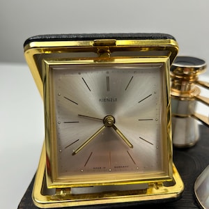 Puede incluir: Un reloj despertador de viaje Kienzle vintage con marco dorado y esfera plateada. El reloj está en un estuche de cuero negro. La esfera del reloj tiene manecillas y marcadores negros. Las palabras "KIENZLE" y "MADE IN GERMANY" son visibles.