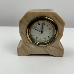 Puede incluir: Pequeño reloj de mármol vintage con bisel dorado y esfera blanca. El reloj tiene números romanos y manecillas negras. El reloj es beige y tiene una base rectangular.