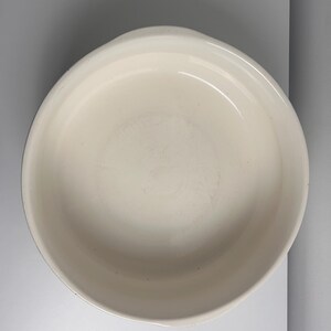 May include: A round, off-white ceramic baking dish with a smooth, glossy interior surface and a wide, slightly raised rim. The vessel features a simple, circular design with integrated side handles, providing a clean and functional appearance.