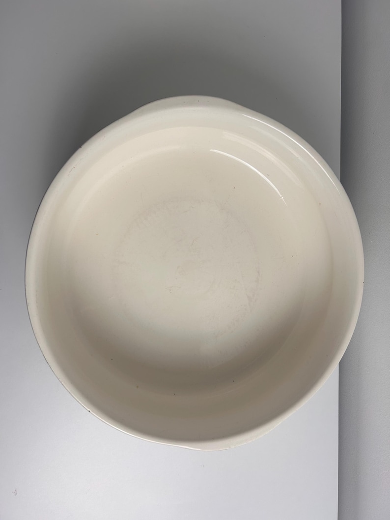 May include: A round, off-white ceramic baking dish with a smooth, glossy interior surface and a wide, slightly raised rim. The vessel features a simple, circular design with integrated side handles, providing a clean and functional appearance.
