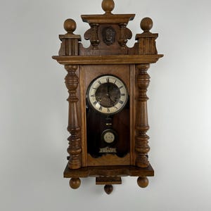 Puede incluir: Un reloj de pared de madera vintage con una esfera redonda y un péndulo. El reloj tiene tallas ornamentadas y elementos decorativos, incluyendo una cara tallada en la parte superior. El reloj es de madera oscura y tiene un diseño clásico y antiguo.