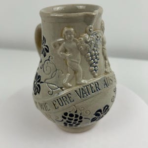 Pode incluir: Jarro pequeno de cerâmica cinza claro com alça e bico. O jarro apresenta querubins em relevo, cachos de uvas e o texto "WIE EURE VATER AUS'S" em uma faixa. Desenhos florais azuis escuros decoram as laterais.