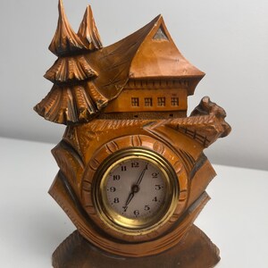 May include: A carved wooden clock shaped like a house with a pine tree on the left side. The clock face is round with a gold-colored metal rim, white background, black numbers from 1 to 12, and two black hands indicating the current time.