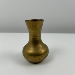 Puede incluir: Un pequeño jarrón de latón con cuerpo redondeado y parte superior acampanada. El jarrón tiene un aspecto ligeramente envejecido, antiguo.