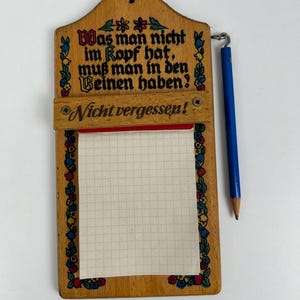 May include: Wooden memo board with a floral border and the German text "Was man nicht im Kopf hat, muss man in den Beinen haben? Nicht vergessen!" A blue pencil hangs from a metal loop. The notepad has a grid pattern.