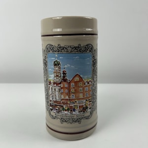 Può includere: Una boccale di birra in ceramica con un design marrone e bianco. La boccale presenta un'immagine di un edificio con una torre dell'orologio e il testo "Künstlerkneipe Striese".