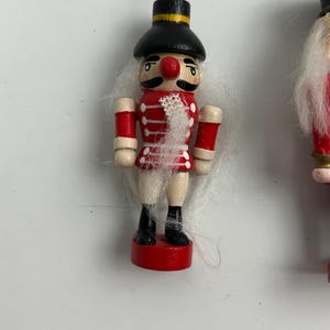 May include: A wooden Christmas ornament shaped like a toy soldier nutcracker. The ornament has a red jacket, black boots, and a black hat with a gold band. The soldier has a white mustache and a red nose. The ornament is suspended by a gold string.