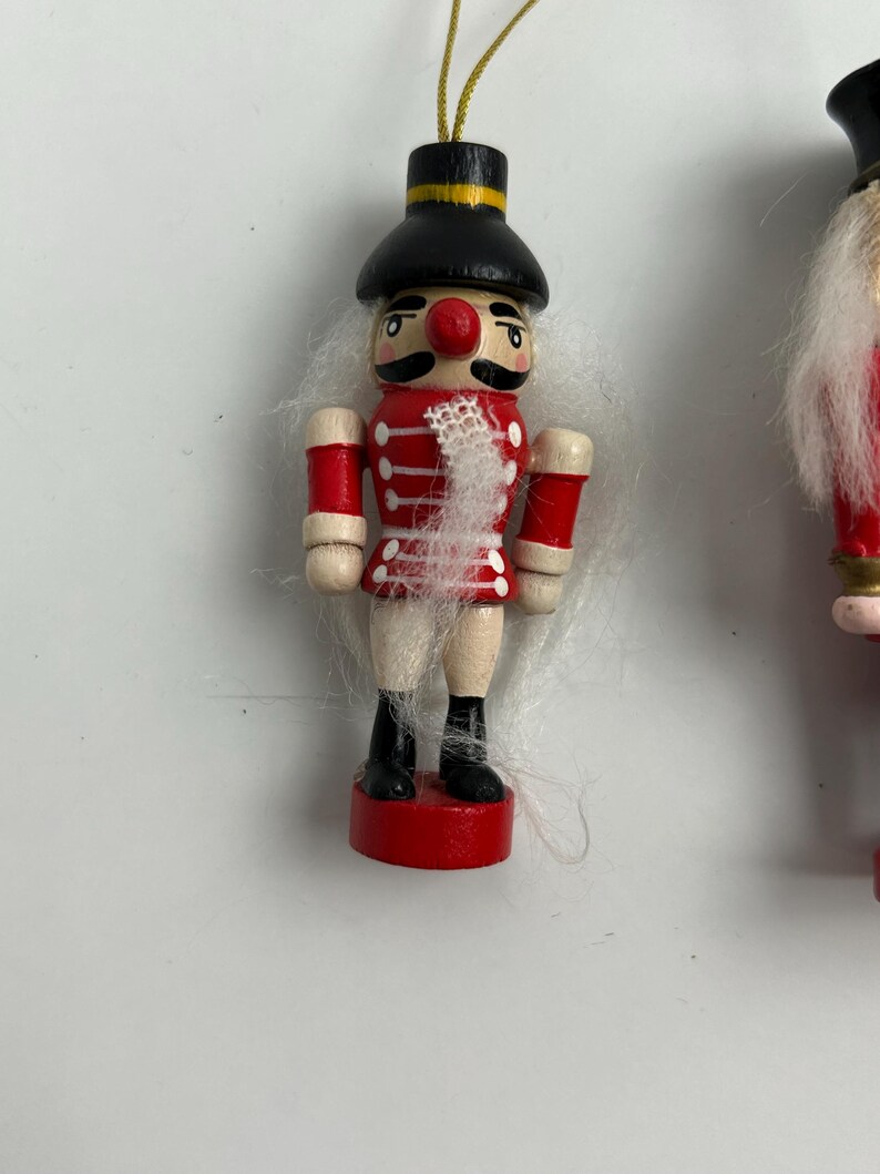May include: A wooden Christmas ornament shaped like a toy soldier nutcracker. The ornament has a red jacket, black boots, and a black hat with a gold band. The soldier has a white mustache and a red nose. The ornament is suspended by a gold string.