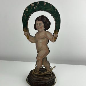 May include: A vintage wooden cherub figurine. The cherub holds a green wreath with gold accents. The cherub is nude with brown hair and stands on a dark wooden base wrapped with twine. The figurine is painted with muted colors.