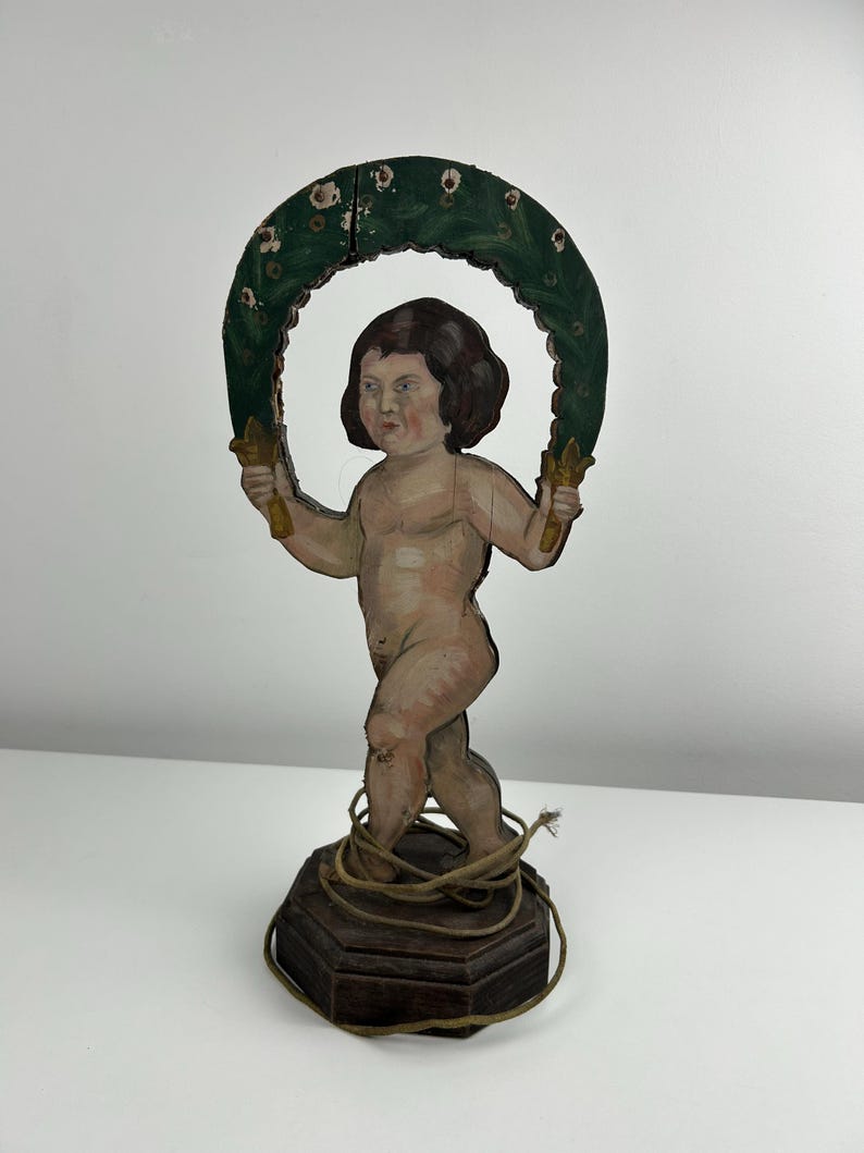 May include: A vintage wooden cherub figurine. The cherub holds a green wreath with gold accents. The cherub is nude with brown hair and stands on a dark wooden base wrapped with twine. The figurine is painted with muted colors.
