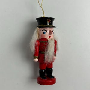 May include: A wooden Christmas ornament in the shape of a nutcracker soldier. The ornament is painted red with black boots and a black hat. It has white hair and a gold string for hanging.