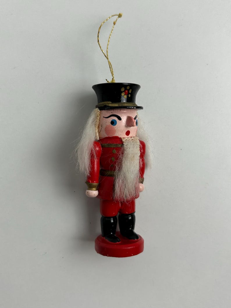 May include: A wooden Christmas ornament in the shape of a nutcracker soldier. The ornament is painted red with black boots and a black hat. It has white hair and a gold string for hanging.