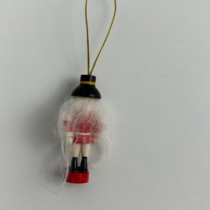 May include: A small, wooden Christmas ornament shaped like a soldier. The ornament features a red jacket, black boots, and a white beard. A gold string is attached for hanging.