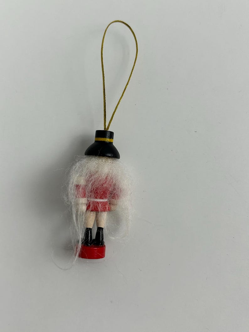 May include: A small, wooden Christmas ornament shaped like a soldier. The ornament features a red jacket, black boots, and a white beard. A gold string is attached for hanging.