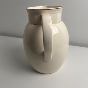 May include: A cream-coloured ceramic jug with a smooth, glossy finish and a rounded body. The jug features a vertical handle on the side and a gold-toned rim around the top opening, providing a simple and functional design for pouring liquids.