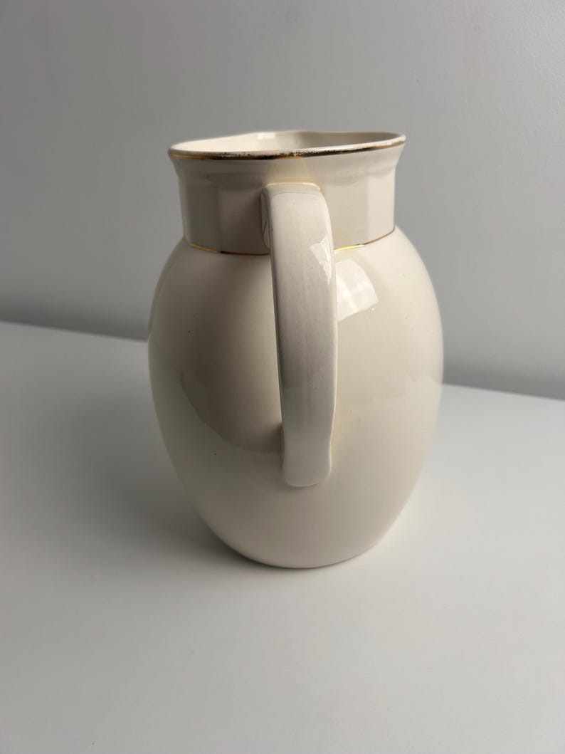 May include: A cream-coloured ceramic jug with a smooth, glossy finish and a rounded body. The jug features a vertical handle on the side and a gold-toned rim around the top opening, providing a simple and functional design for pouring liquids.