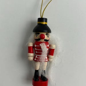 May include: A wooden Christmas ornament in the shape of a toy soldier. The ornament has a black hat with a yellow band, a red coat with white accents, and black boots. The soldier has a red nose and white hair. A gold string is attached for hanging.