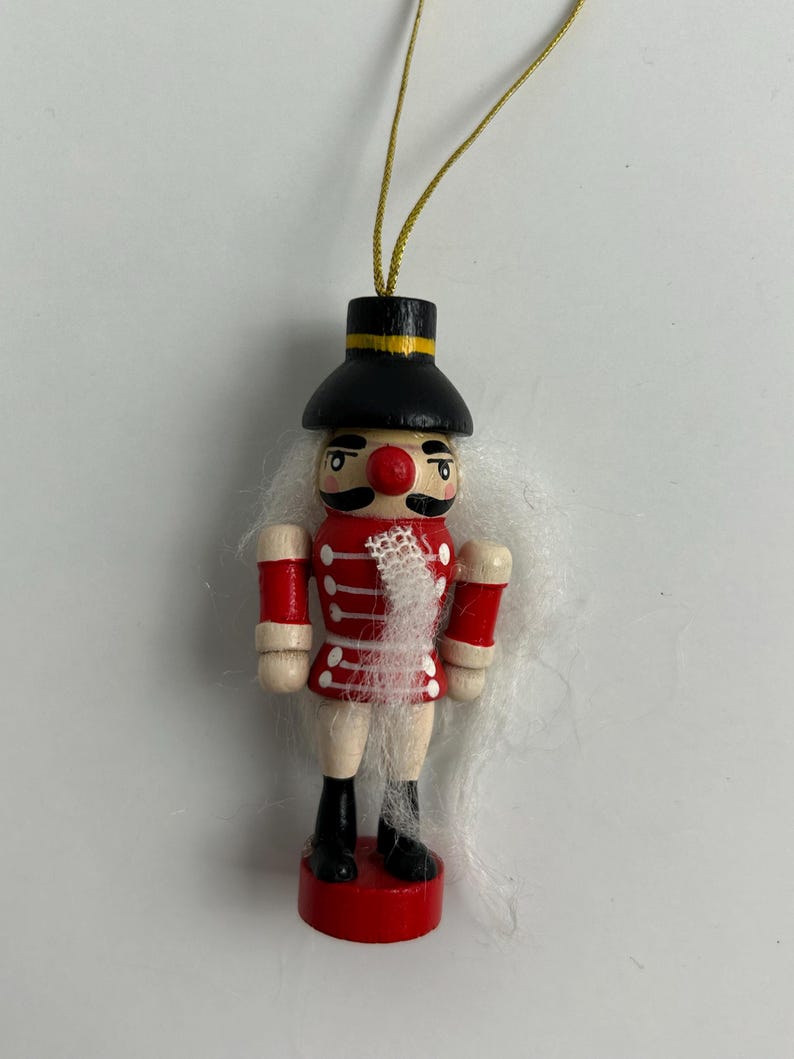 May include: A wooden Christmas ornament in the shape of a toy soldier. The ornament has a black hat with a yellow band, a red coat with white accents, and black boots. The soldier has a red nose and white hair. A gold string is attached for hanging.