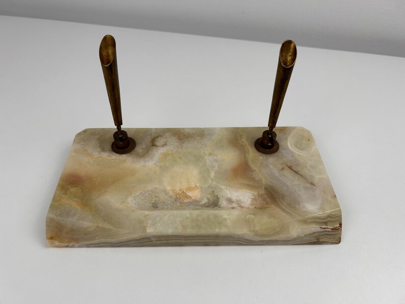 Op de afbeelding: Een rechthoekige pennenhouder van lichtgekleurde steen met bruine en groene adering. Twee messing penhouders zijn aan de steen bevestigd. De penhouders zijn omhoog gericht. De pennenhouder is een bureau-accessoire.