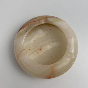 Op de afbeelding: Een ronde asbak van natuursteen met een gladde, gepolijste afwerking. De asbak is voornamelijk cr&egrave;mekleurig met bruine en oranje strepen. Het heeft een ronde kom en een kleine inkeping voor sigaretten.