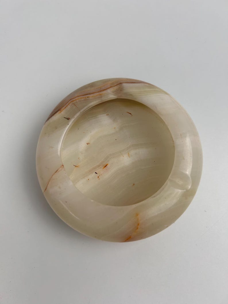 Op de afbeelding: Een ronde asbak van natuursteen met een gladde, gepolijste afwerking. De asbak is voornamelijk cr&egrave;mekleurig met bruine en oranje strepen. Het heeft een ronde kom en een kleine inkeping voor sigaretten.