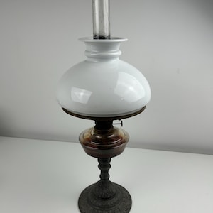 May include: An antique oil lamp with a white glass shade and a clear glass chimney. The lamp has a dark metal base and a decorative, ornate design. The oil reservoir is a dark amber color.