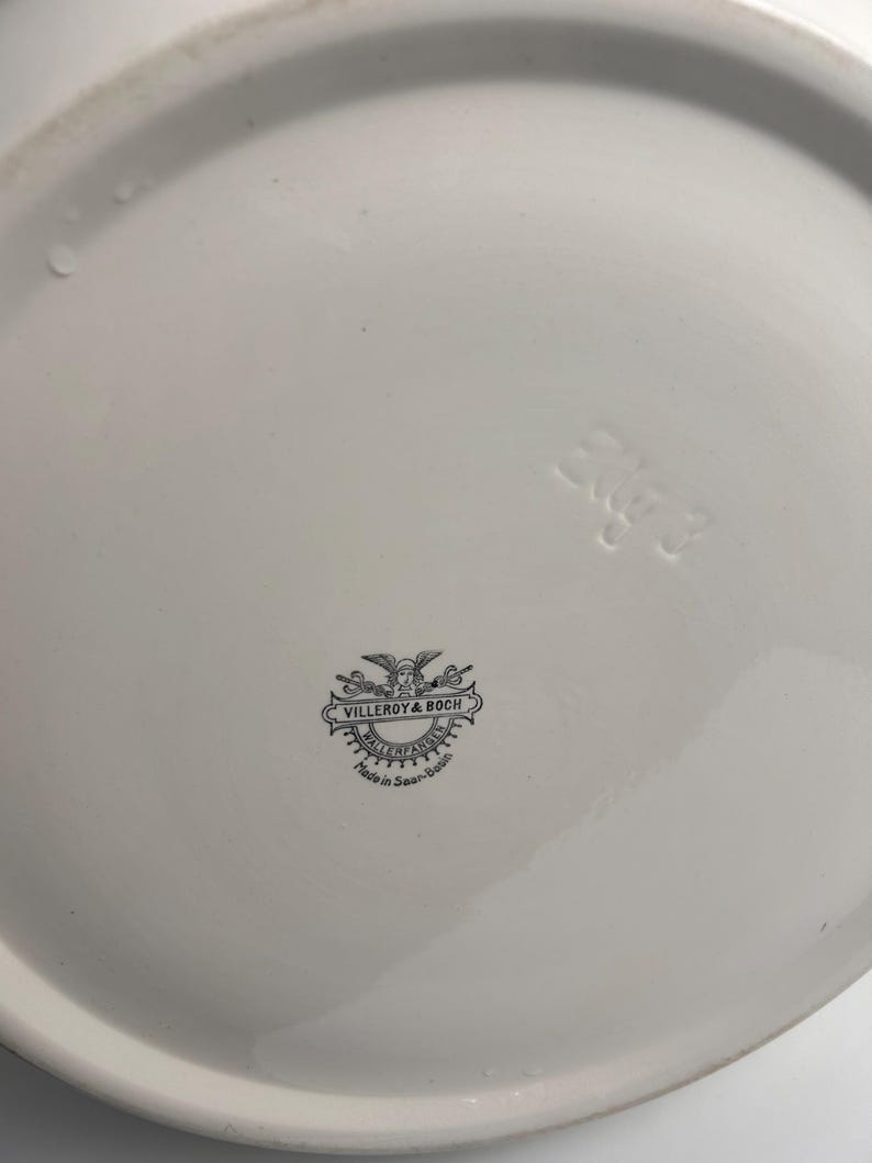 May include: The underside of a white ceramic plate featuring a black printed Villeroy & Boch logo with a winged figure and the text Wallerfangen Made in Saar-Basin. An embossed number 3 is visible on the smooth, glossy white surface.
