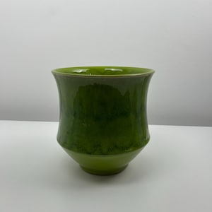 May include: A green ceramic vase with a flared top and a rounded base. The vase has a glossy finish and a gradient of green hues, from a lighter shade at the base to a darker shade at the top. The vase is a decorative item.