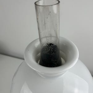 May include: A white, round vase with a wide, circular opening and a clear glass tube inserted in the center. The vase has a glossy finish and a dark interior visible through the glass tube.