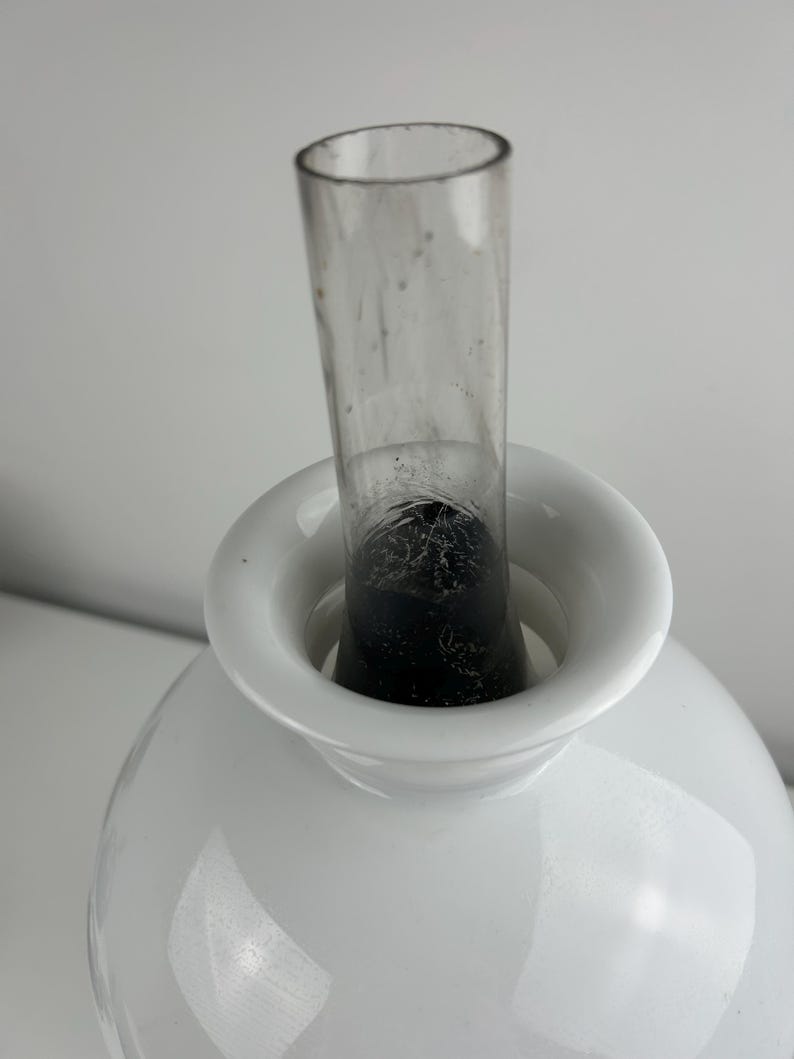 May include: A white, round vase with a wide, circular opening and a clear glass tube inserted in the center. The vase has a glossy finish and a dark interior visible through the glass tube.